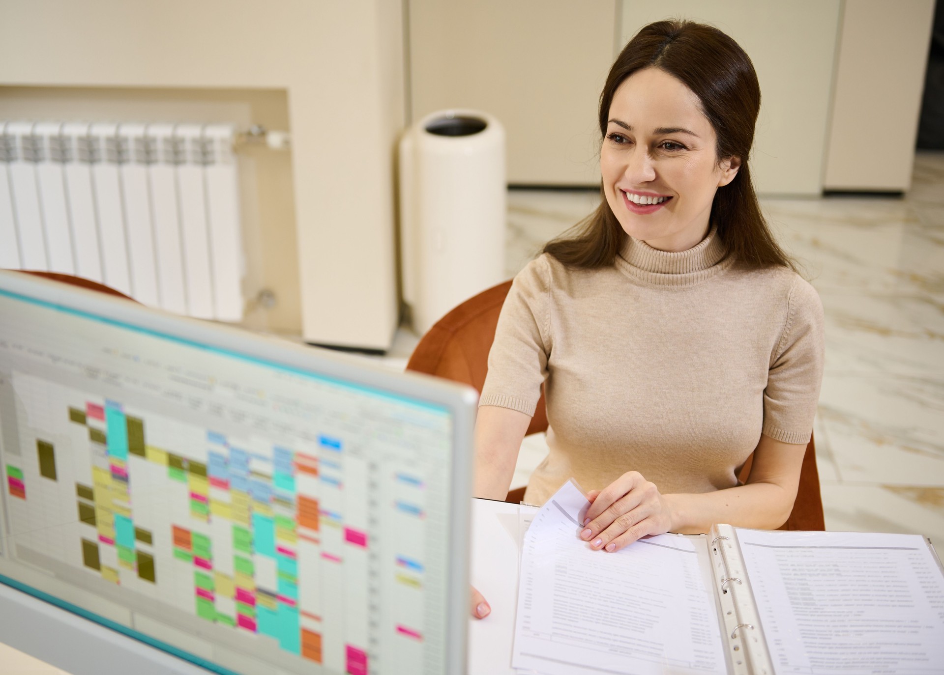 Attractive woman browsing the price-list of consumer services sitting at reception desk of a modern office with switched on monitor on the foreground